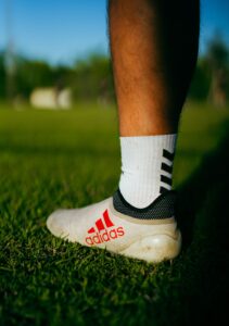 7 July - Agencies – African Marketing Confederation – African Marketing Confederation Close-up of a white Adidas soccer boot on green grass, with a white sock and red Adidas logo visible at the heel area.