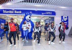 Group of people dancing inside a Stanbic Bank lobby, with a large blue bear mascot and bank signage visible in the background.