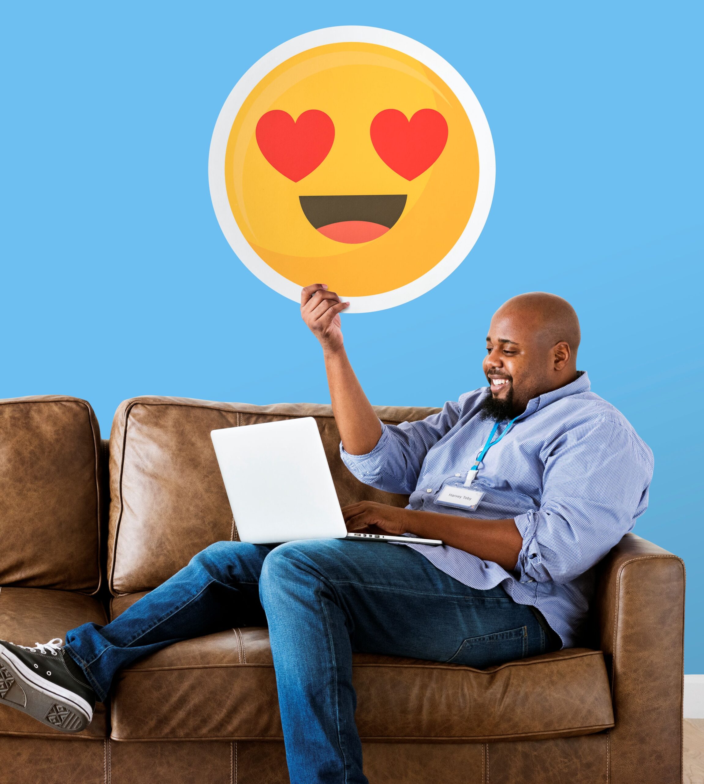 Man sitting on a brown leather sofa holding a large round emoji with heart-shaped eyes against a blue background.