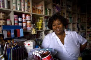 Smiling woman behind a cluttered cosmetics counter with shelves of lotions, creams, and makeup jars behind her.