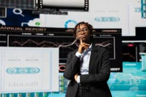 Professional woman in a blazer stands with hand on chin in front of large data dashboards and charts in a high-tech control room.