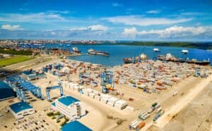 Aerial view of a busy shipping port with stacked containers, blue gantry cranes, and ships along the harbor under a blue sky.