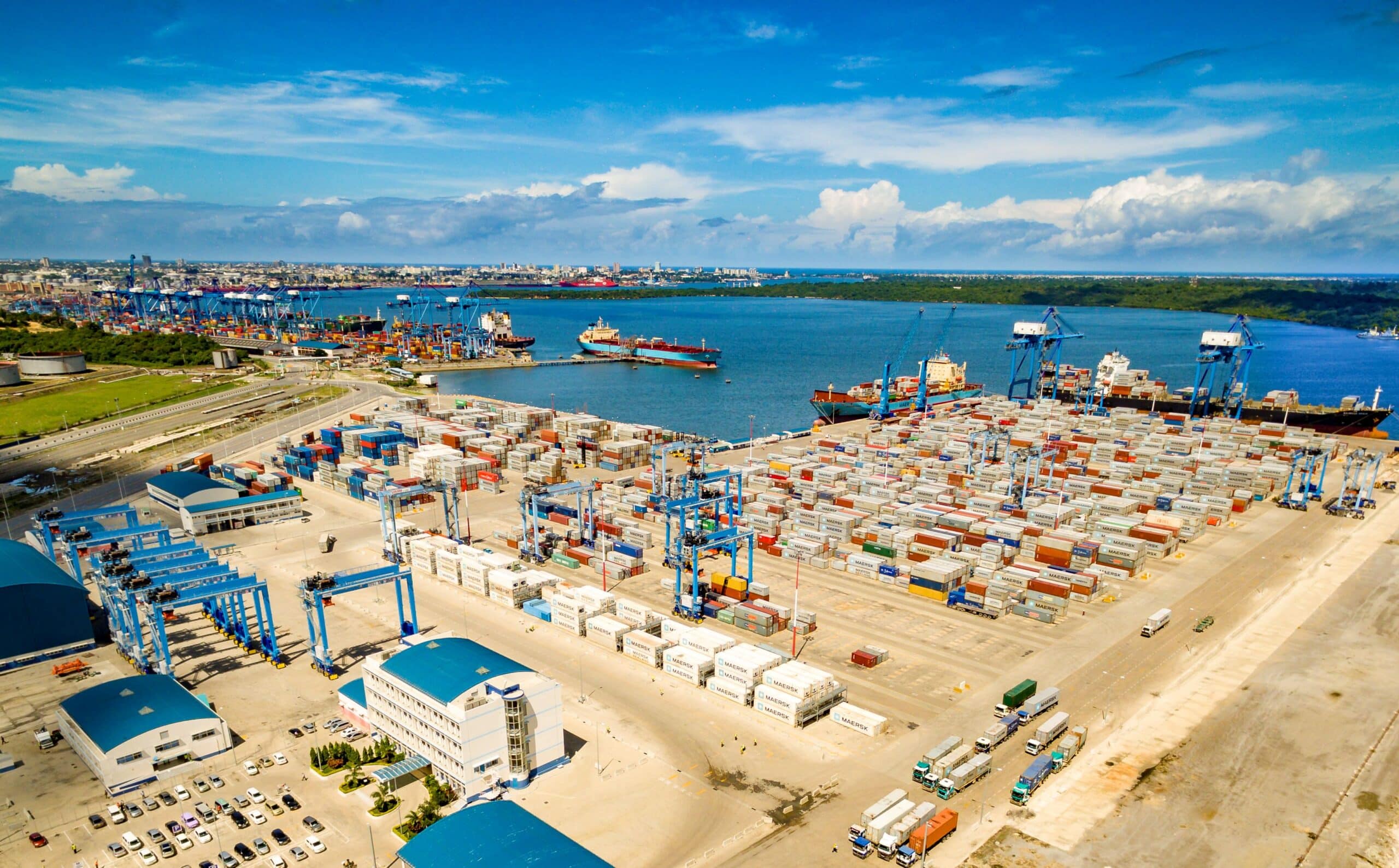 Aerial view of a busy shipping port with stacked containers, blue gantry cranes, and ships along the harbor under a blue sky.