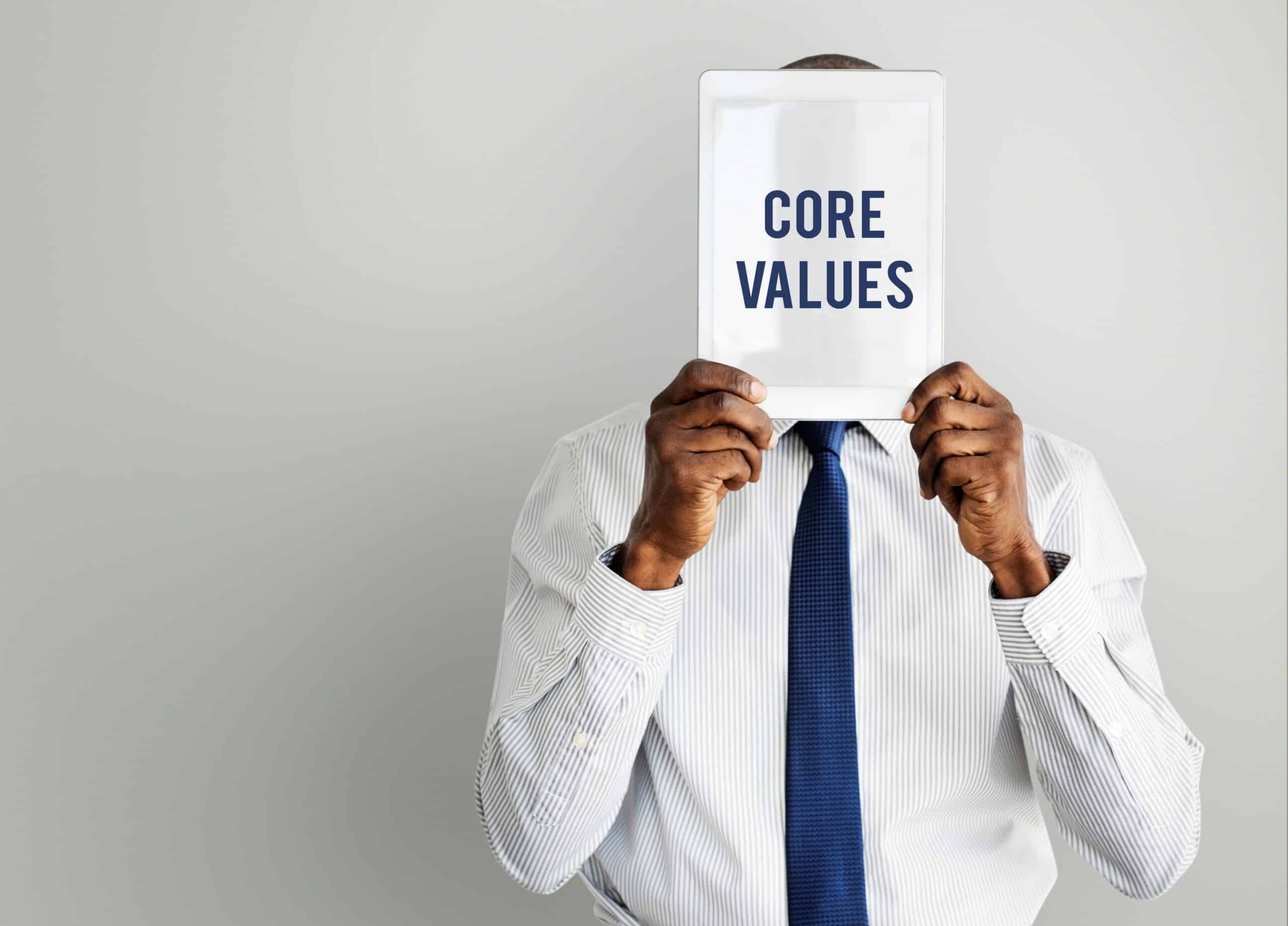 Person in a striped shirt and blue tie holds up a sign obscuring their face that reads CORE VALUES in bold letters.