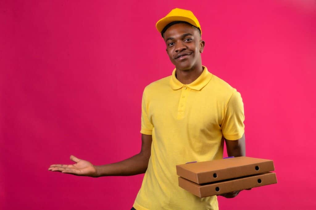 Delivery worker in a yellow polo and cap holds three stacked cardboard boxes against a bright pink background.