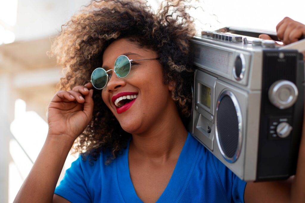 Smiling woman with curly hair and round sunglasses stands beside a vintage boombox resting on her shoulder.