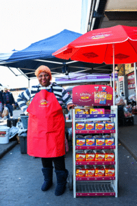 August Route to Market Market vendor in a red albany apron stands beside a wheeled display rack of albany dairy products under red and blue umbrellas