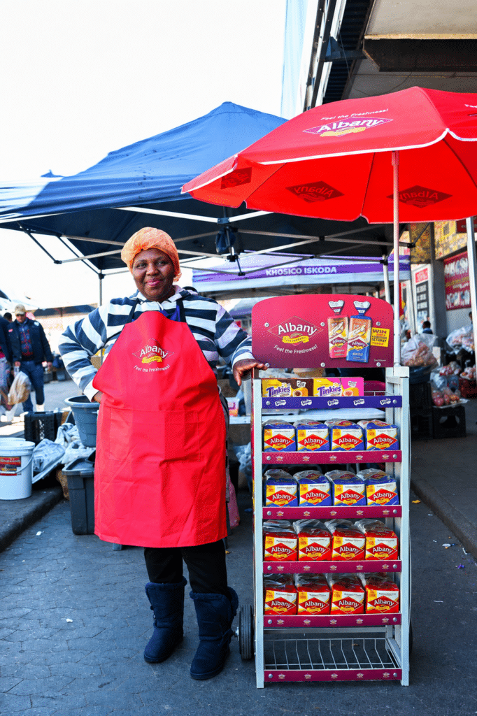 Market vendor in a red Albany apron stands beside a wheeled display rack of Albany dairy products under red and blue umbrellas.