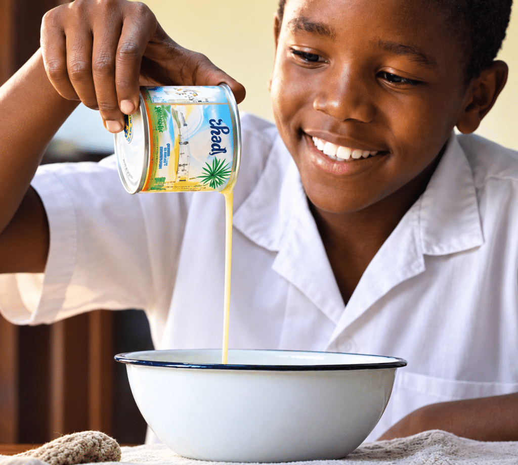 Smiling boy in a white shirt pours a thick yellow liquid from a tropical-design can into a large white bowl.