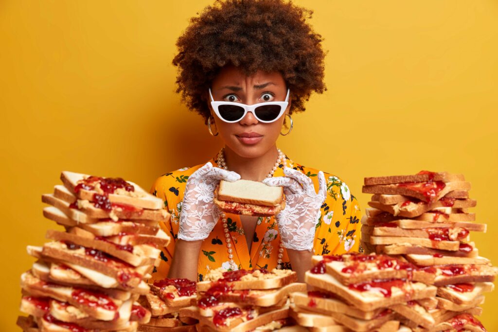Woman with an Afro, white sunglasses, and pearl necklace holds tall stacks of peanut butter and jam sandwiches against a yellow background.
