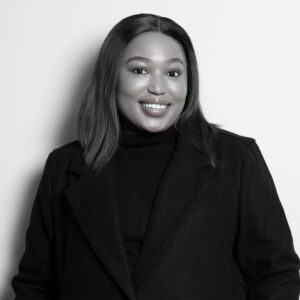 Smiling woman of color in a black coat poses against a light background, looking at the camera.