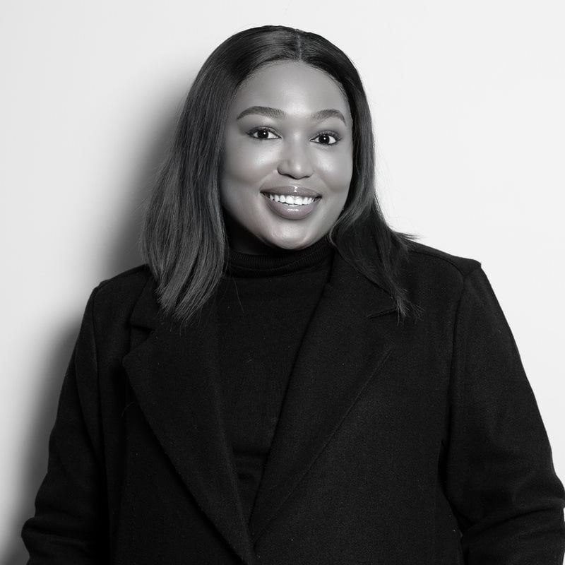 Smiling woman of color in a black coat poses against a light background, looking at the camera.