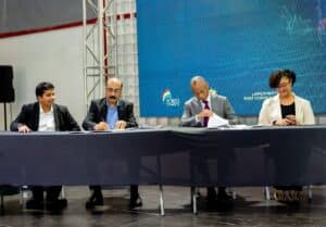 Panel of four professionals seated at a long table, signing documents, with a blue digital backdrop displaying a Porto News logo behind them.