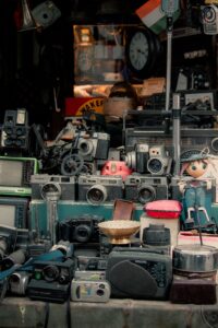 Cluttered display of vintage cameras and electronics, including Polaroid and Canon models, on a shelf with a doll figure on the right.