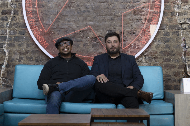 Two men sit on a blue couch against a brick wall, with a large circular neon sign behind them, casually posing together.