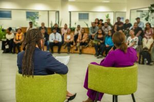 Two presenters sit in lime-green chairs with their backs to the camera, addressing a diverse audience in a bright lobby.