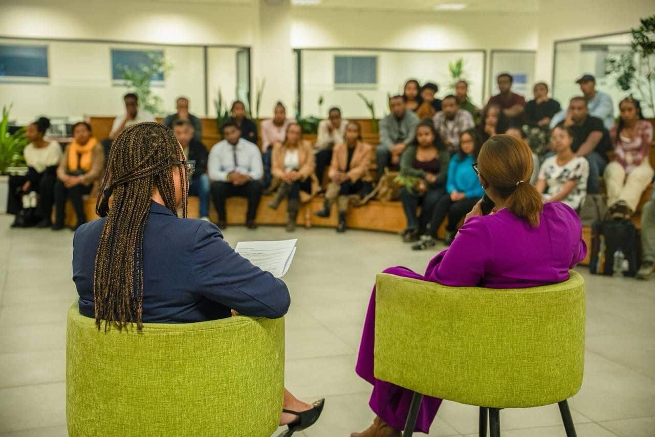 Two presenters sit in lime-green chairs with their backs to the camera, addressing a diverse audience in a bright lobby.