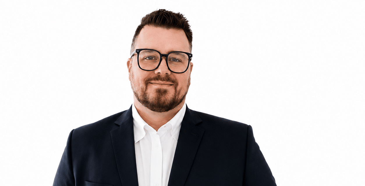 Professional headshot: man in a navy suit and white shirt, wearing glasses, smiling at the camera.