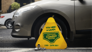 Yellow triangular sign in front of a parked silver car promoting Paddy Power Comedy Festival (23–26 July).