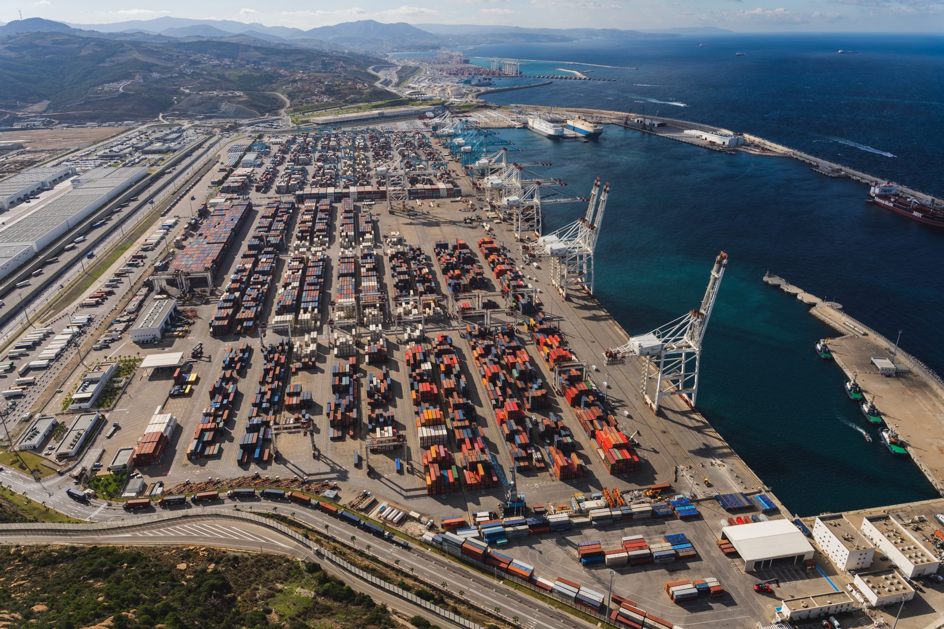 6 August - Supply Chain – African Marketing Confederation – African Marketing Confederation Aerial view of a busy container port with rows of stacked cargo containers, large cranes, and ships along the dock.