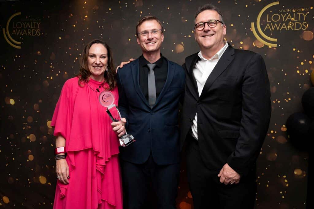 Three people in formal attire pose for a photo at an awards event; a woman in a pink dress holds a trophy while two men in suits stand beside her against a gold-confetti backdrop.