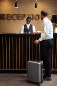 September Tourism Marketing Hotel receptionist assisting a guest at the check in counter with a gray suitcase nearby behind the desk