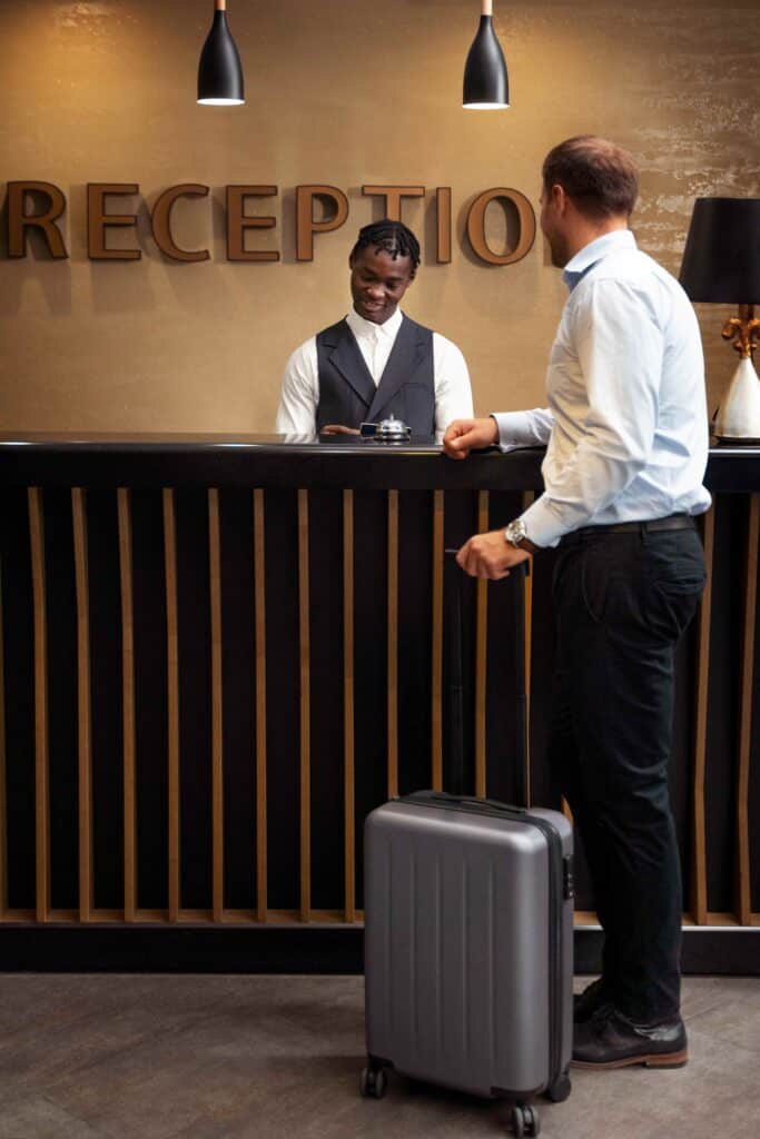 Hotel receptionist assisting a guest at the check-in counter with a gray suitcase nearby behind the desk.