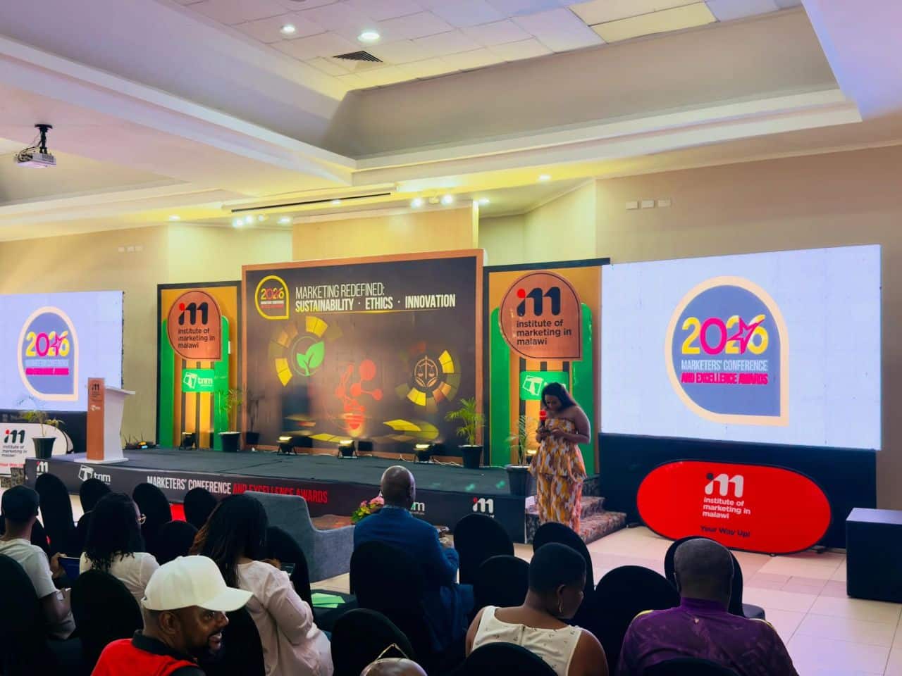 September Amc Member News Conference stage with a woman in a patterned dress speaking to an audience large screens display '' logos and banners for a marketers conference with 'institute of marketing in malawi' branding on stage panels and backdrop