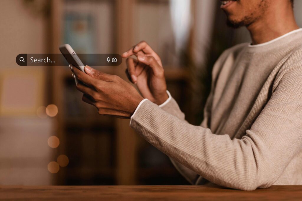 Person wearing a beige sweater holds a phone with a translucent search bar reading 'Search…' and microphone/camera icons in the foreground.