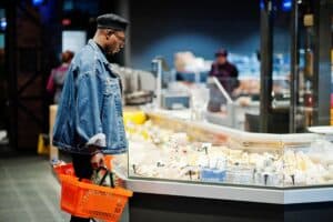 Person shopping at a deli counter holding an orange shopping basket in a cheese section of a supermarket