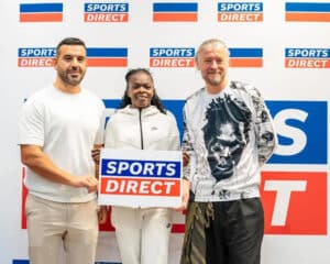 Three people stand side by side in front of a sports direct backdrop the woman in the middle holds a blue and orange 'sports direct' sign