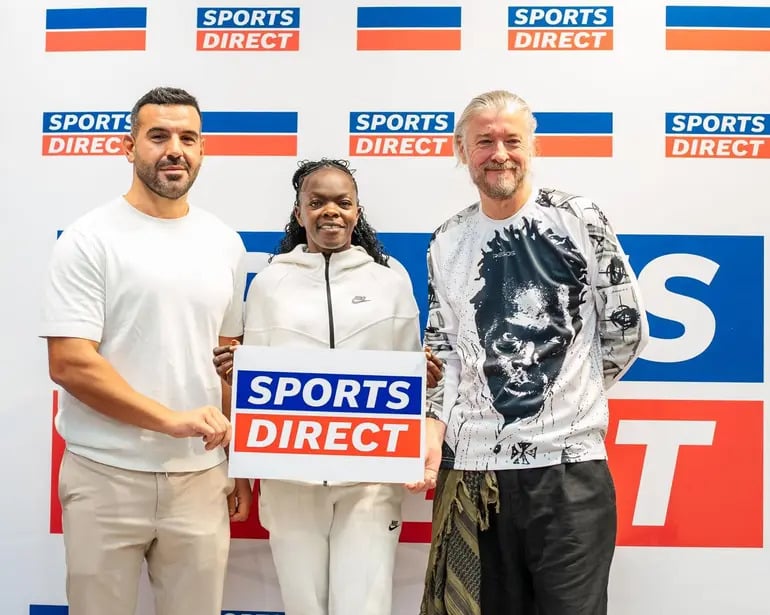 Three people stand side by side in front of a sports direct backdrop the woman in the middle holds a blue and orange &apossports direct&apos sign