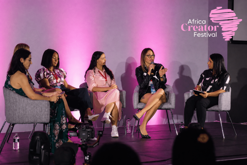 Panel of five women seated on stage at Africa Creator Festival, speaking into mics under pink lighting, logo visible in top right.