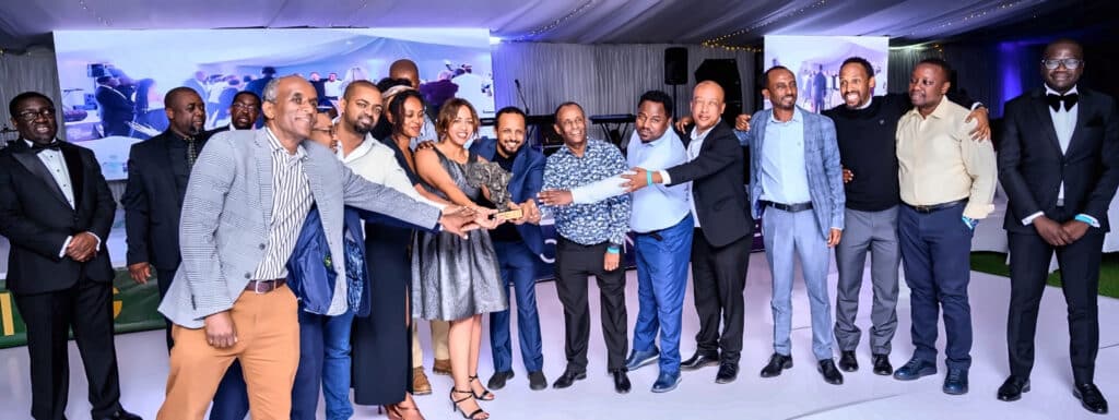 Group of people in formal attire posing together, reaching toward a central trophy at an awards event on a white runway stage with a blue backdrop.