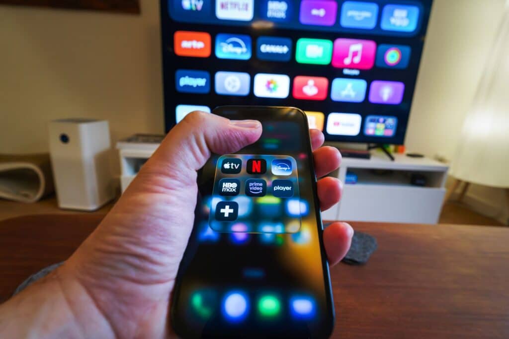 Person holding two smartphones with app icons visible on their screens; a large TV screen in the background shows colorful app icons.