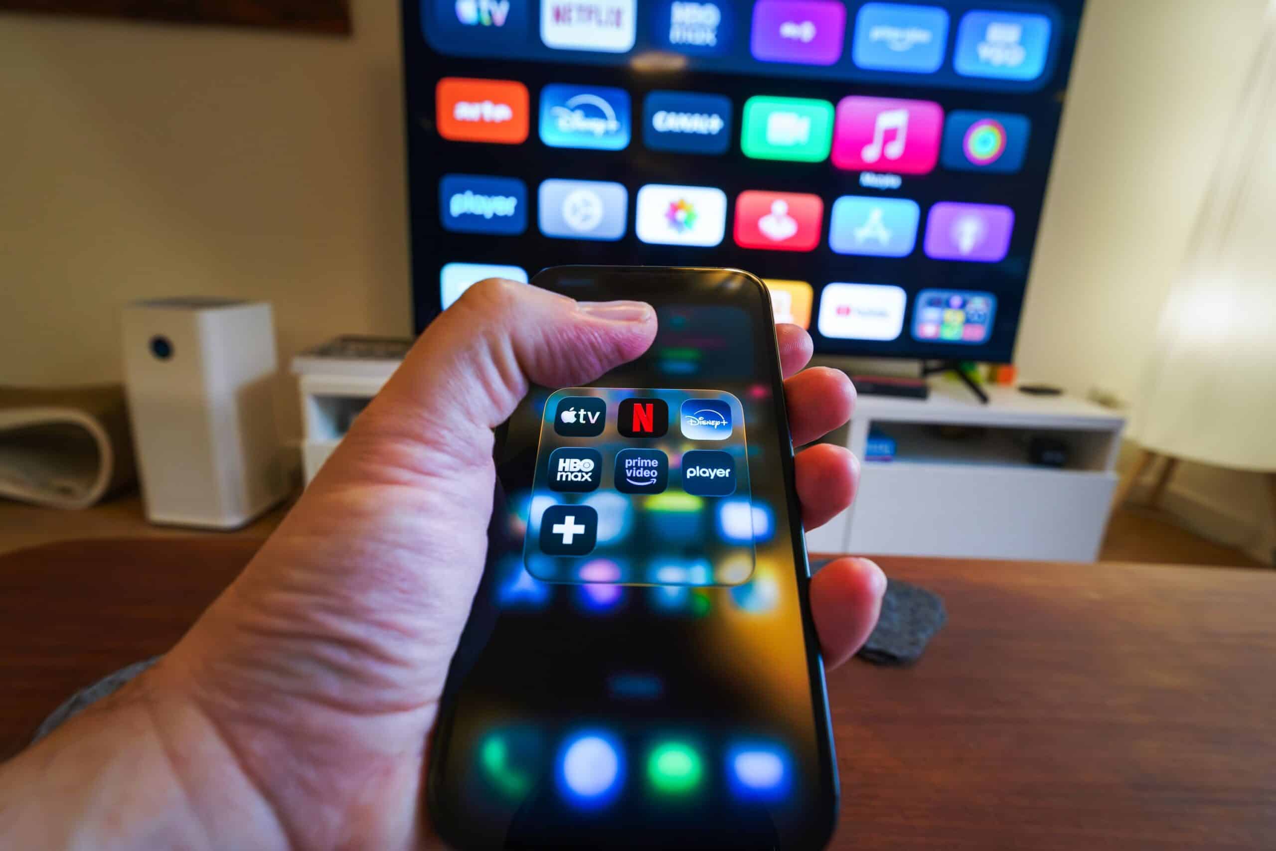 Person holding two smartphones with app icons visible on their screens a large tv screen in the background shows colorful app icons