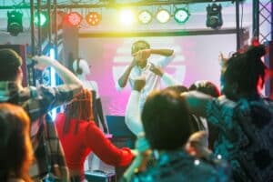 Dj performing on a brightly lit stage as colorful lights shine over a dancing crowd