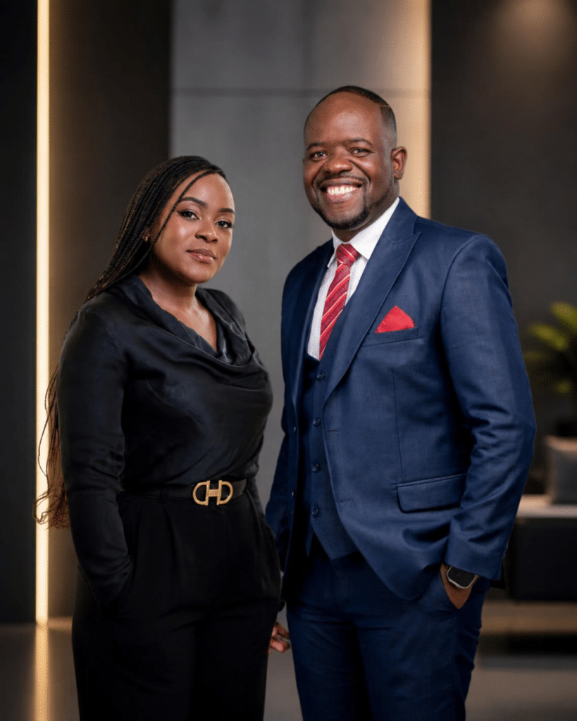 Two professionals posing together in a modern office lobby; man in a blue suit with red tie and pocket square, woman in black outfit beside him.
