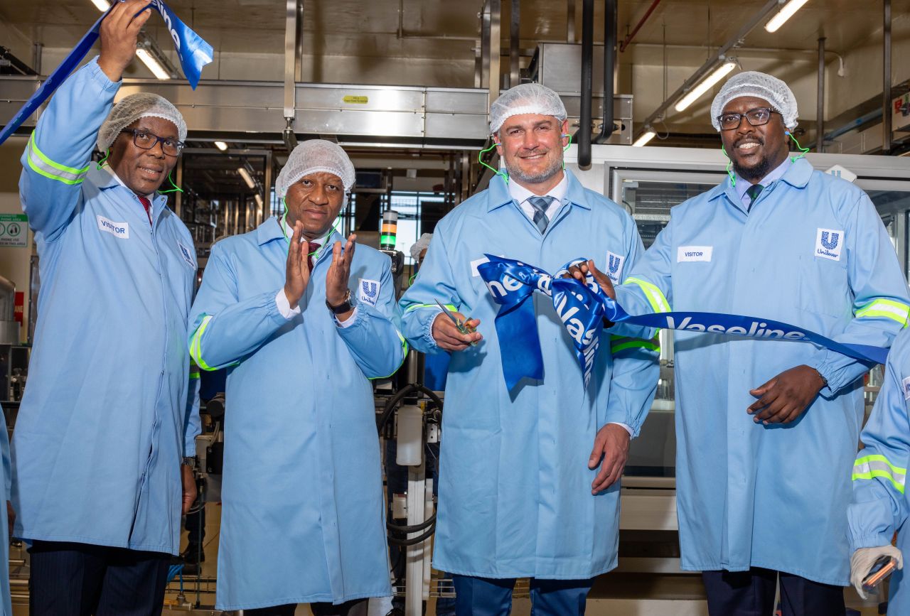 Four professionals in blue protective coats and hairnets cut a blue ribbon during a factory ceremony celebrating a ribbon cutting event