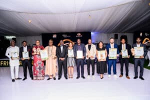 Group of award recipients posing on stage each holding certificates and trophies under a white tent backdrop