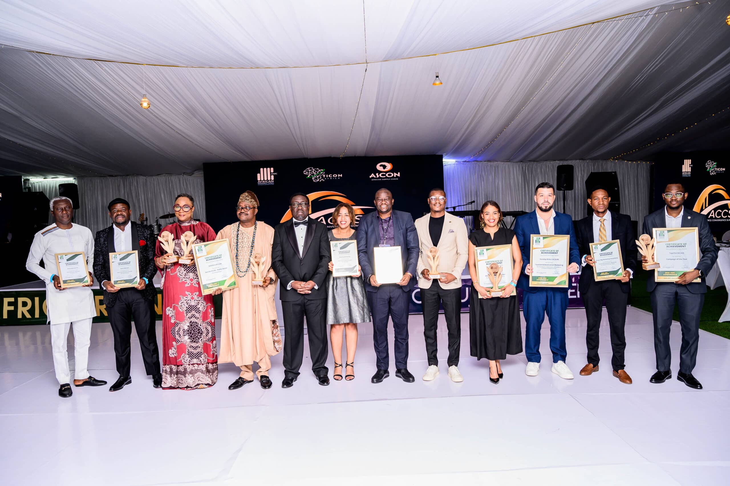 Group of award recipients posing on stage each holding certificates and trophies under a white tent backdrop