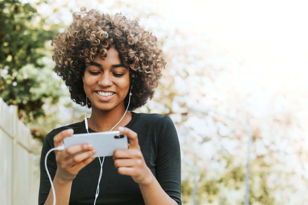 Young woman with curly hair smiling while looking at her phone outdoors, wearing earphones.