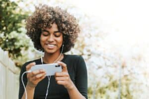 Young woman with curly hair smiling while looking at her phone outdoors wearing earphones