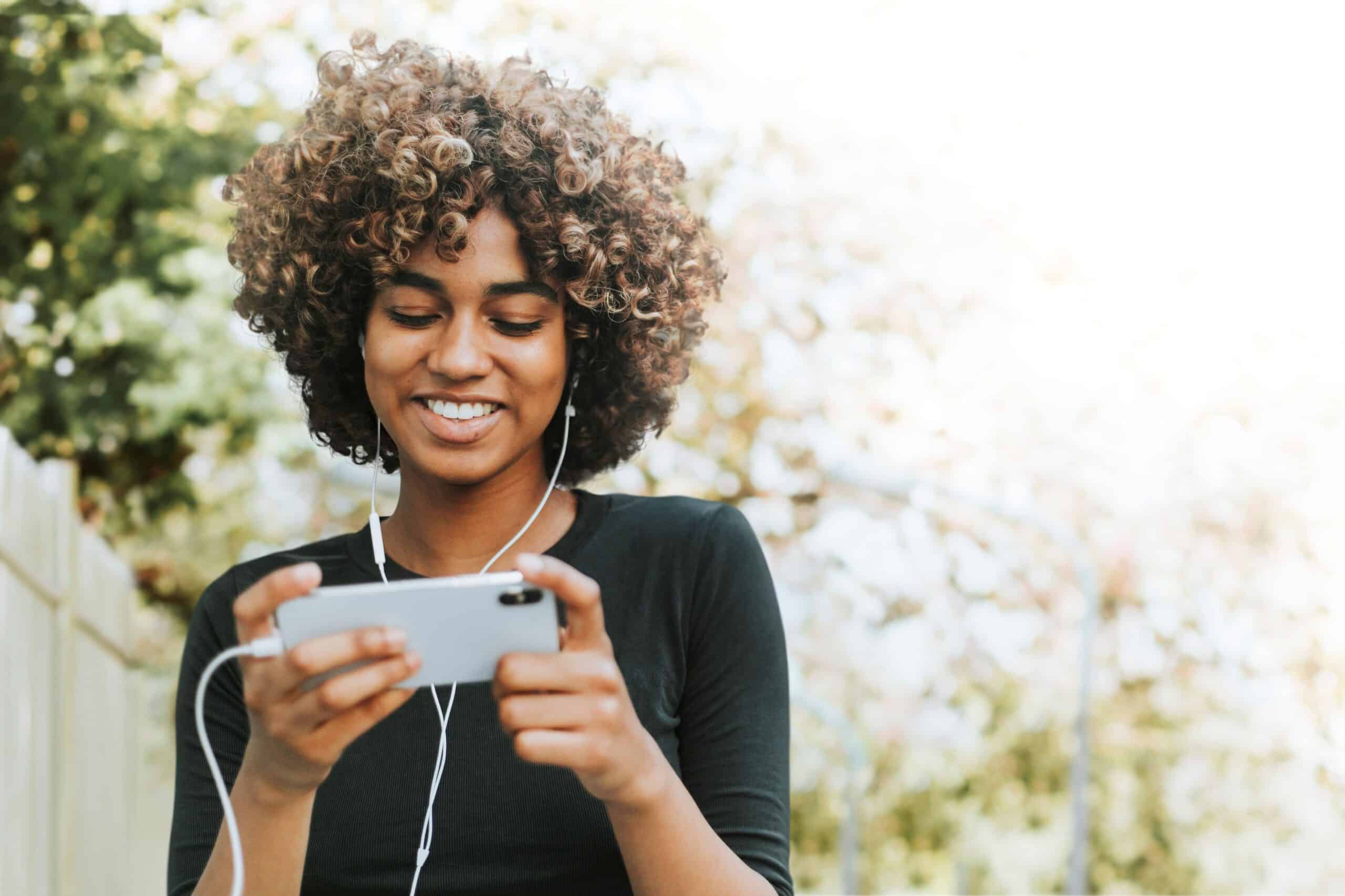 Young woman with curly hair smiling while looking at her phone outdoors wearing earphones