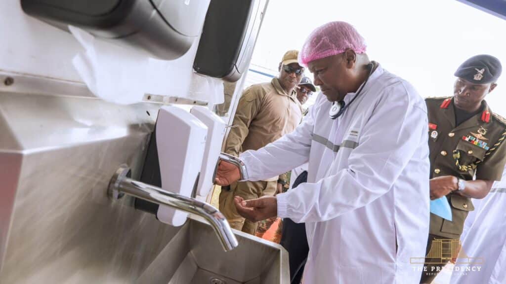 Medical professional in a white coat and pink hairnet demonstrates handwashing at a stainless steel station while soldiers watch nearby.