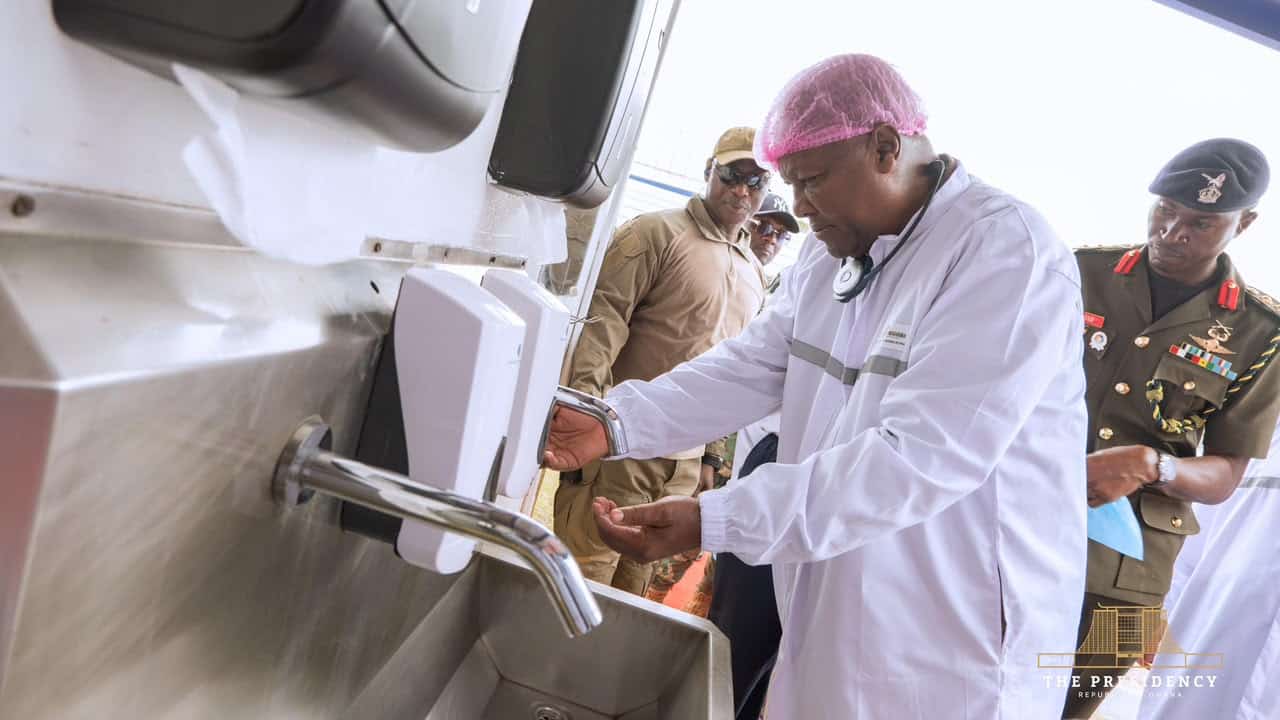 Medical professional in a white coat and pink hairnet demonstrates handwashing at a stainless steel station while soldiers watch nearby