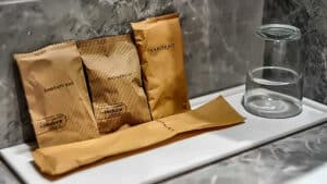 Gold hotel toiletries sachets arranged on a marble bathroom counter including a vanity kit and shower gel beside a glass