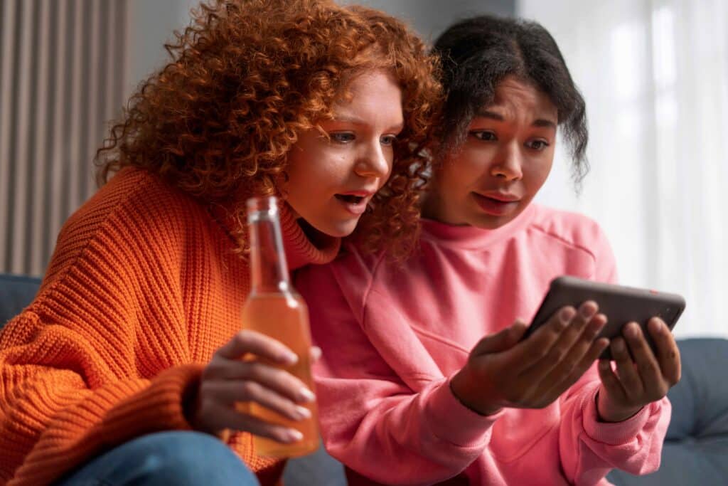 Two young women sit together, looking at a tablet; one in an orange sweater holds a bottle while the other in a pink hoodie eyes the screen.