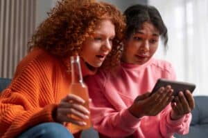 October Advertising Research Two young women sit together looking at a tablet one in an orange sweater holds a bottle while the other in a pink hoodie eyes the screen