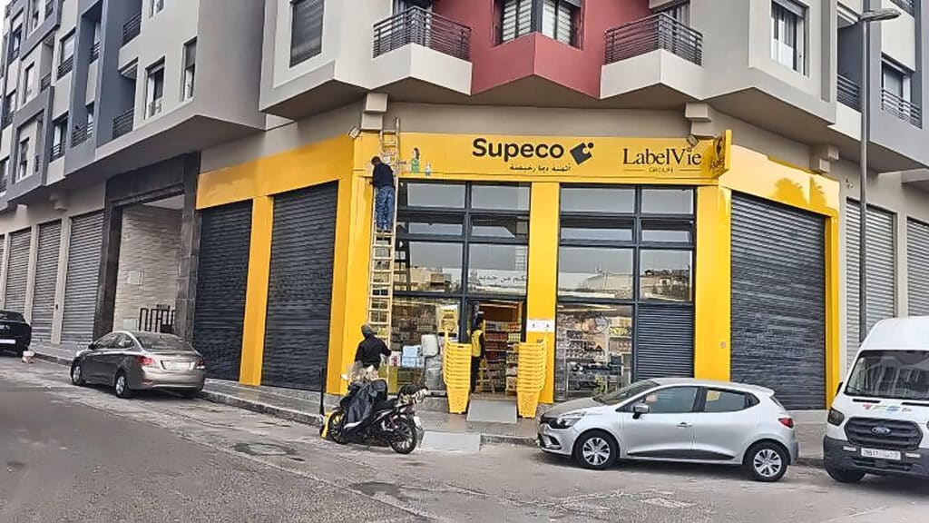 Worker on a ladder painting the yellow storefront of a corner shop labeled Supeco and Label Vie, with shoppers and parked vehicles nearby.